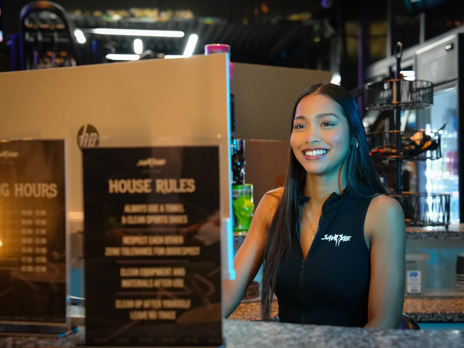 Savage Elite Fitness front desk host smiling beside the house rules board at reception